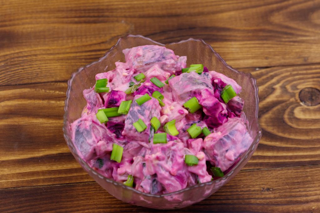 Klassischer Heringssalat mit Rote Bete und Ei in einer Glasschale, garniert mit frischen Frühlingszwiebeln, auf einem Holztisch serviert.
