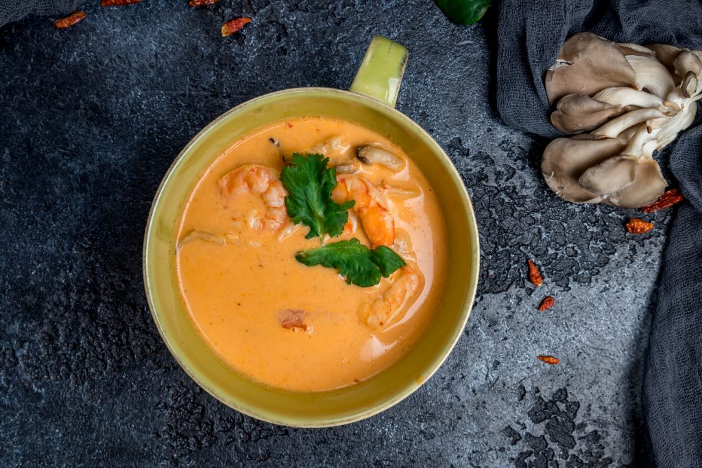 Authentische Tom Yam Suppe in einer Schale, mit Garnelen, Pilzen und frischen Kräutern, serviert auf dunklem Hintergrund.