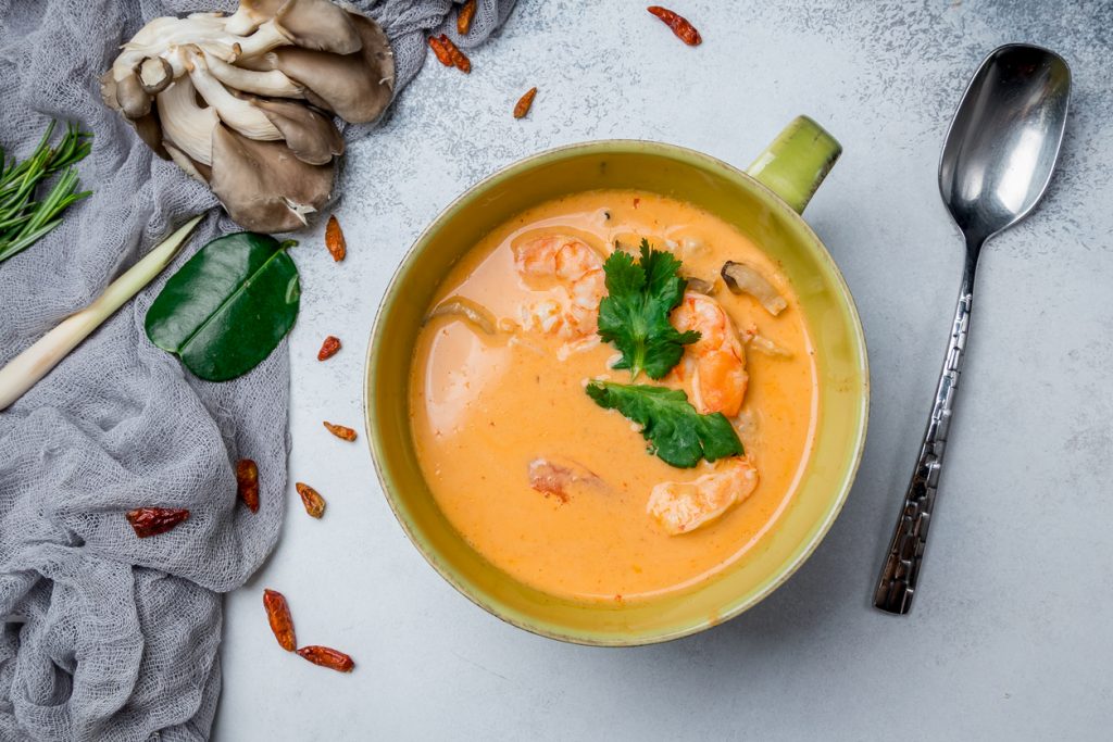 Thailändische Tom Yam Suppe mit Garnelen, Pilzen und frischen Kräutern, serviert in einer grünen Schale mit Löffel daneben.