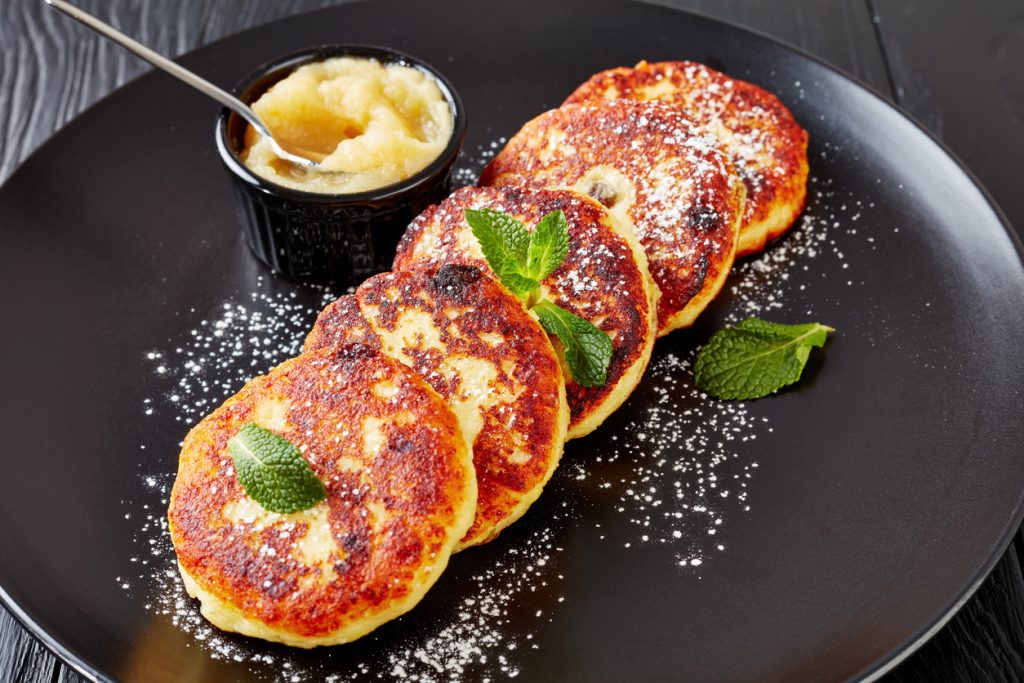 Quark-Kartoffelpfannkuchen auf schwarzem Teller, serviert mit Apfelmus, Minzblättern und Puderzucker bestäubt.