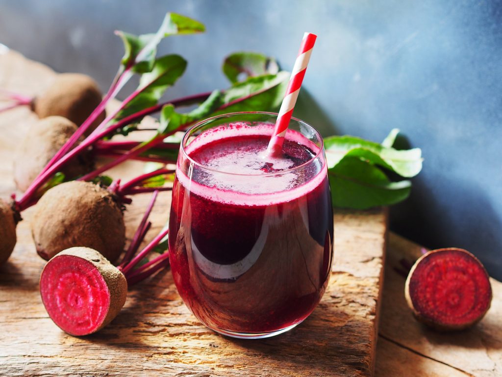 Glas mit frischem Rote-Bete-Saft und Strohhalm, daneben frische rote Beete mit Blättern.