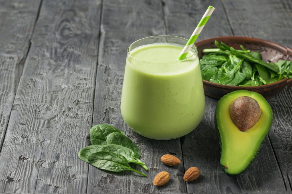 Glas mit cremigem Spinat-Avocado-Smoothie auf Holzoberfläche; frische Zutaten dezent im Hintergrund.