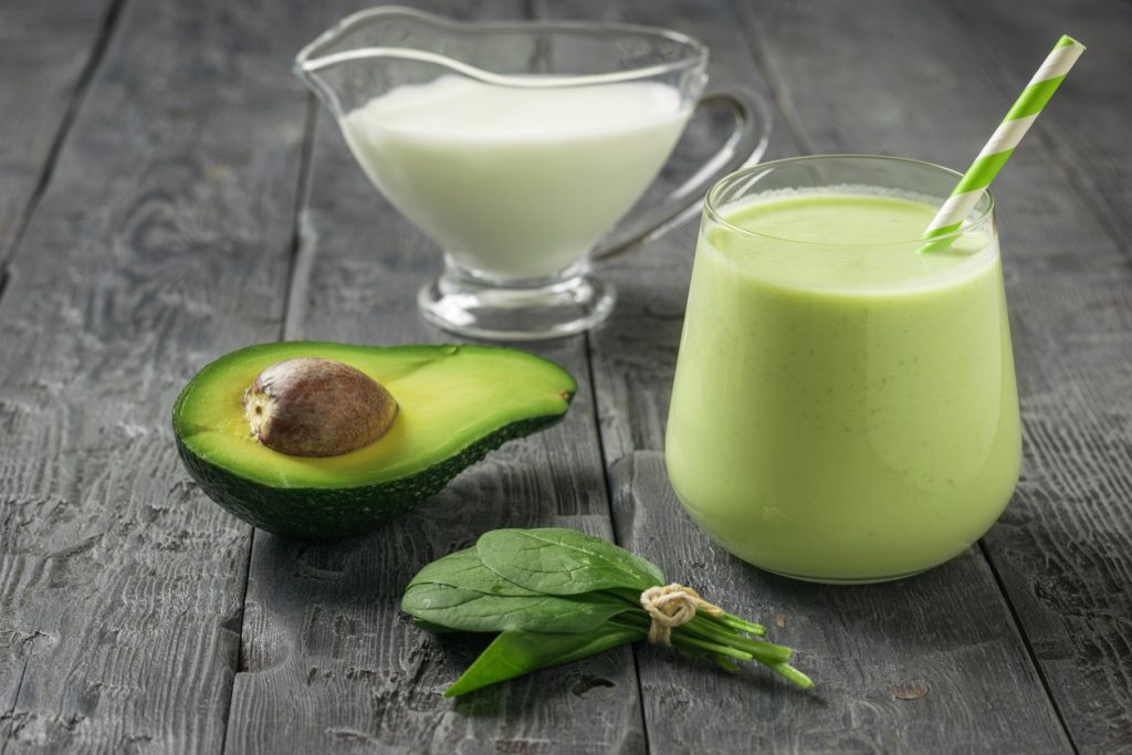 Glas mit grünem Avocado-Spinat-Smoothie; daneben Avocadohälfte, frischer Spinat und Kännchen Mandelmilch.