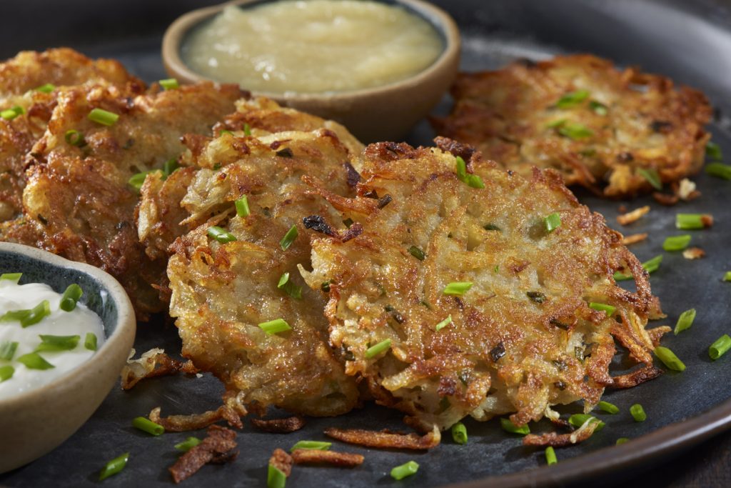 Nahaufnahme von goldbraunen, knusprigen Kartoffelpuffern, mit frischem Schnittlauch bestreut; daneben Schälchen mit Apfelmus und cremigem Dip auf dunklem Teller.