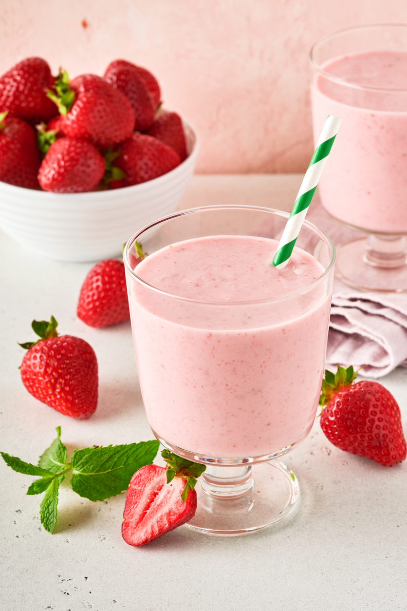 Glas mit rosa Erdbeer-Shake und grün-weißem Strohhalm, daneben halbierte Erdbeeren, Minzblätter und eine Schale voller Früchte vor rosafarbenem Hintergrund.