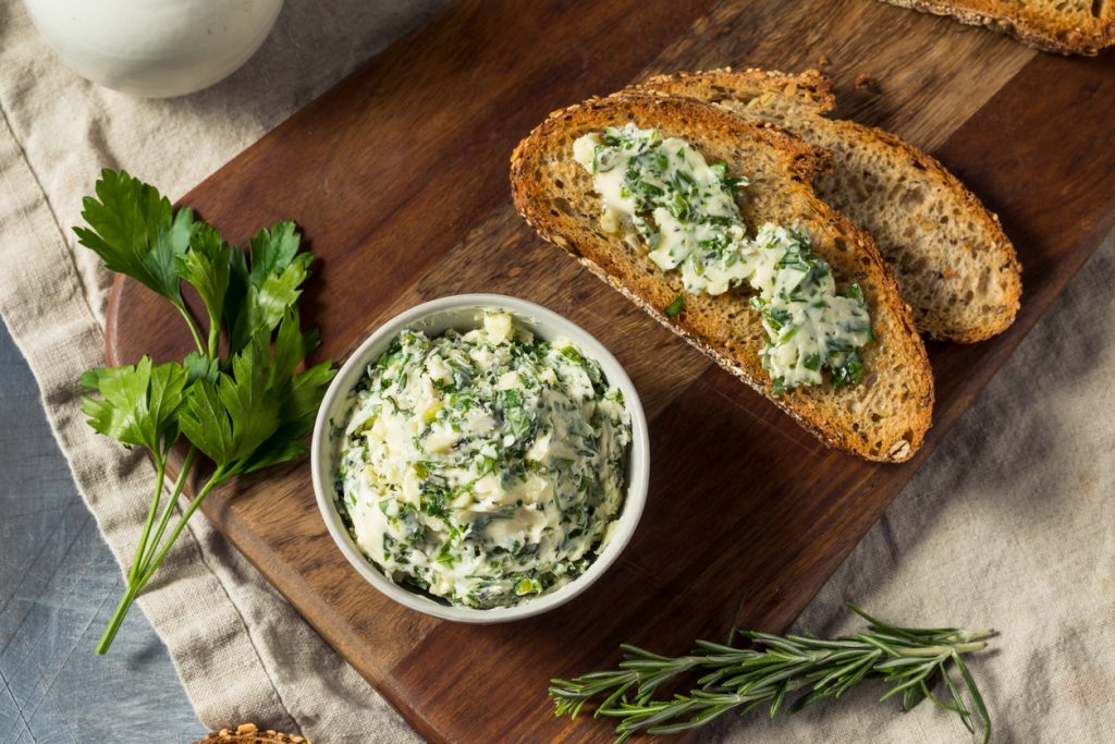 Schälchen Kräuterbutter auf Holzbrett mit bestrichenem Brot, Petersilie und Rosmarin.