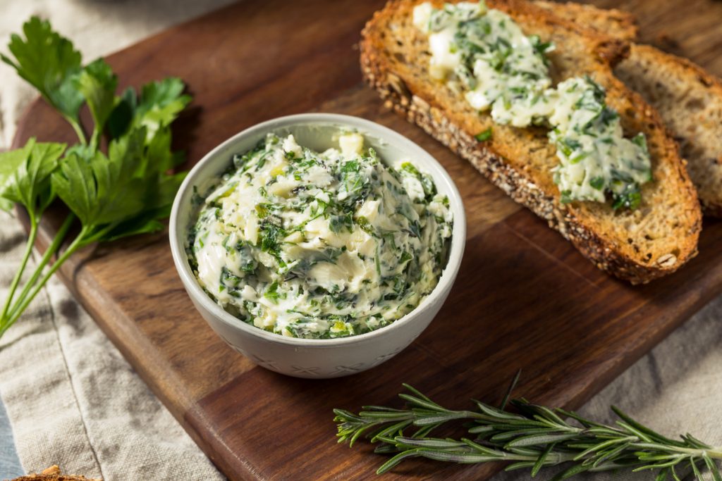 Schälchen Kräuterbutter auf Holzbrett mit Brot, Petersilie und Rosmarin, Draufsicht.