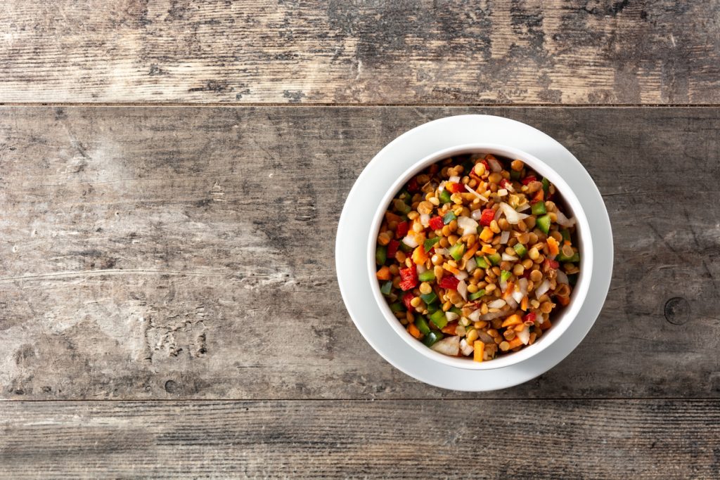 Schüssel mit buntem Linsensalat, Draufsicht auf Holztisch mit großzügiger Leerfläche.