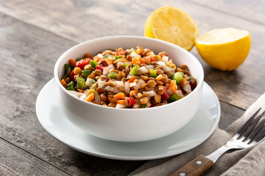 Schüssel mit buntem Linsensalat und Zitronenhälften auf Holztisch, daneben Gabel.