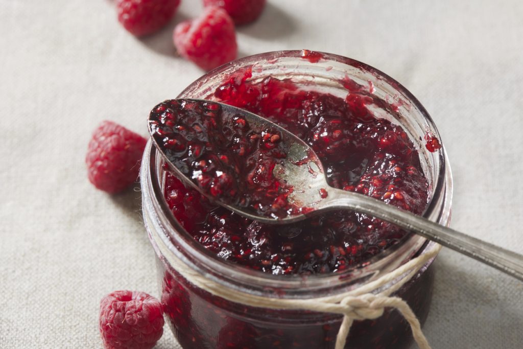 Glas mit Himbeermarmelade, ein Löffel liegt darauf, frische Himbeeren daneben arrangiert.