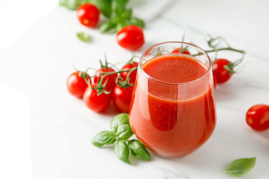 Glas mit frischem Tomatensaft auf weißem Tisch, dekoriert mit Basilikumblättern und ganzen Tomaten an der Rispe.