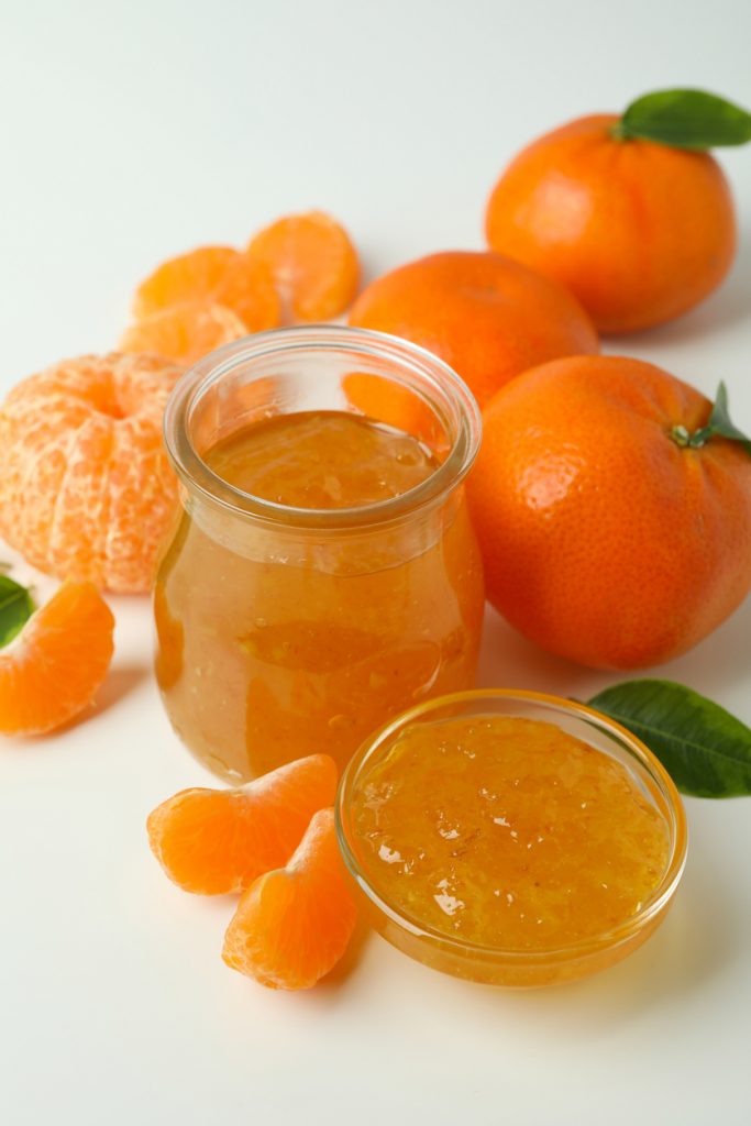 Ein Glas und eine Schale mit leuchtend orangener Mandarinenmarmelade, umgeben von ganzen Mandarinen, geschälten Spalten und grünen Blättern auf weißem Hintergrund.