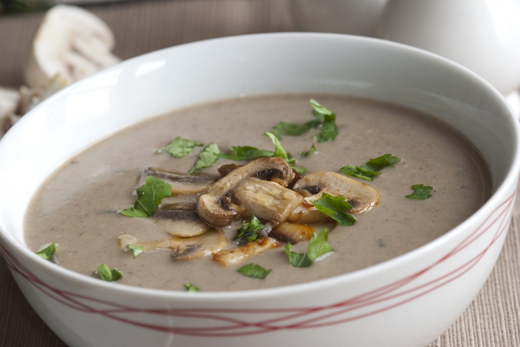 Weiße Schüssel mit cremiger Champignonsuppe, garniert mit frischen Champignonscheiben und gehackter Petersilie.