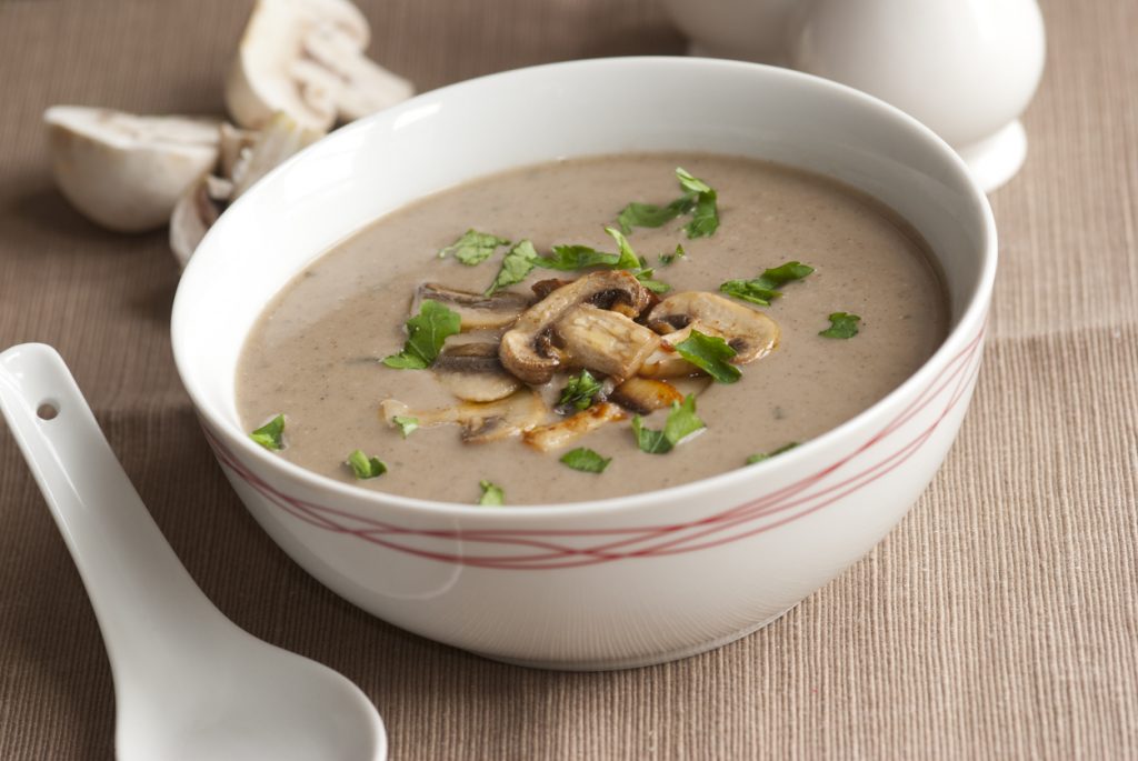 Cremige Champignonsuppe in einer weißen Schüssel, garniert mit gebratenen Champignons und Petersilie, daneben ein Porzellanlöffel.