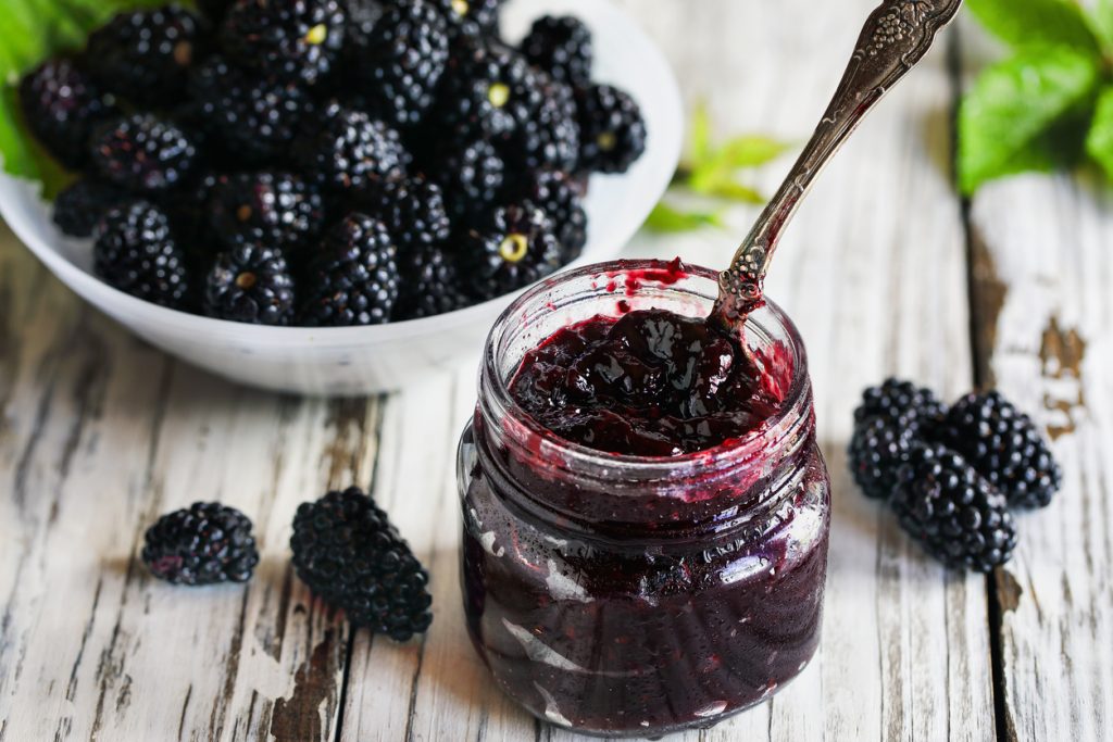 Ein Glas mit selbstgemachter Brombeermarmelade und Löffel, daneben frische Brombeeren in einer Schale auf einem rustikalen Holztisch.