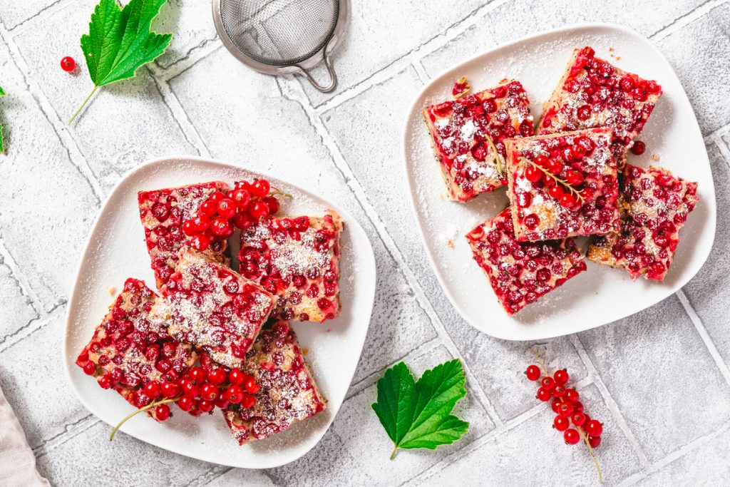 Stücke von Johannisbeer-Blechkuchen mit Mohn, dekoriert mit Puderzucker und frischen Johannisbeeren, auf zwei Tellern angerichtet.
