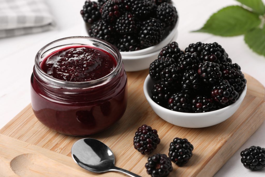 Ein Glas mit selbstgemachter Brombeermarmelade, daneben frische Brombeeren in Schalen auf einem Holzbrett.