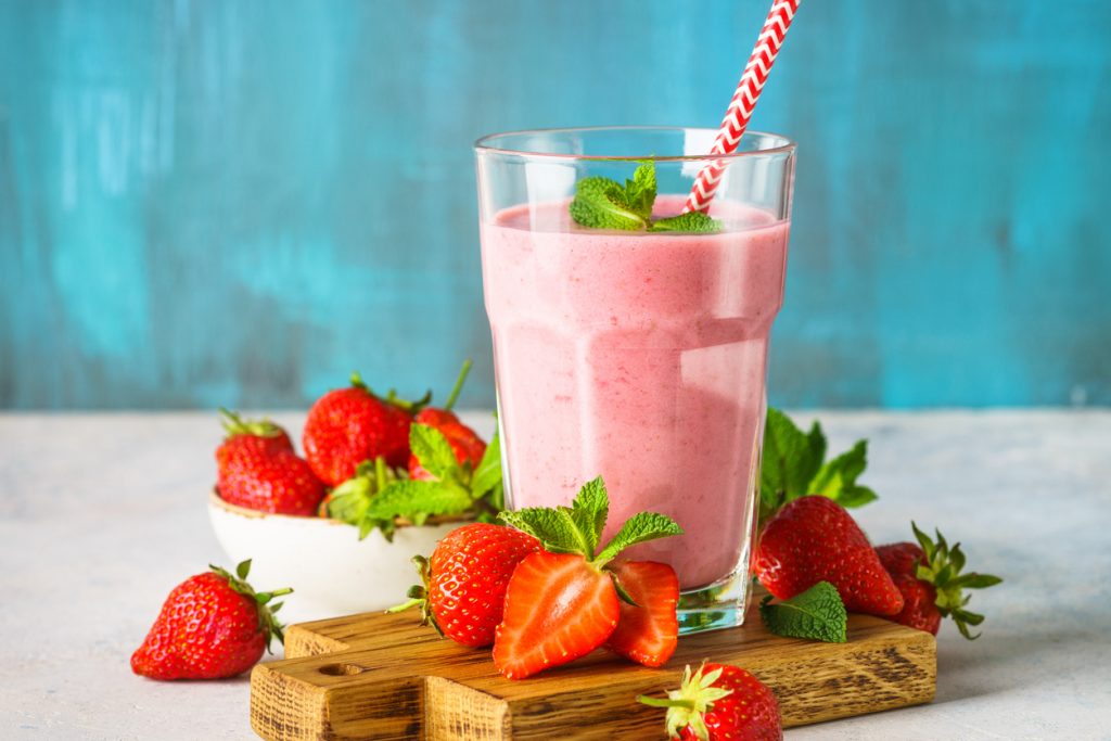 Cremiger Erdbeershake in einem Glas mit rotem Strohhalm, frischen Erdbeeren und Minzblättern auf Holzbrett vor türkisfarbigem Hintergrund.