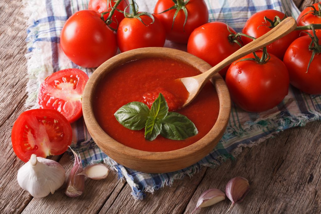 Holzschale mit Tomatensauce und Basilikum, Holzlöffel; frische Tomaten und Knoblauch auf Holztisch