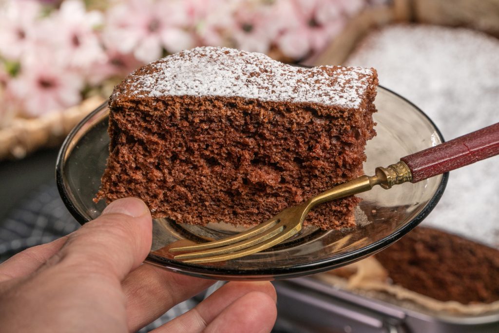 Stück Schokoladenkuchen mit Puderzucker auf Glasteller, goldene Gabel, Hand hält den Teller