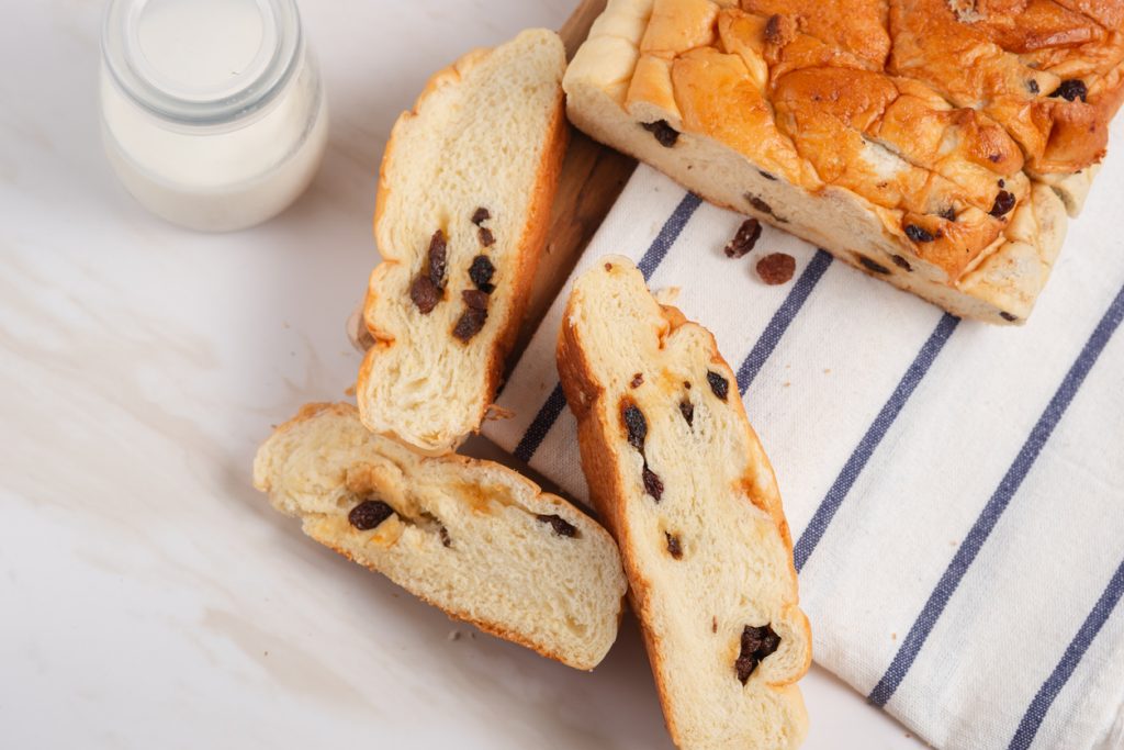 Aufgeschnittenes frisches Rosinenbrot auf einem gestreiften Tuch, daneben eine Flasche Milch.