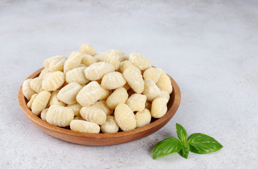 Schale mit frischen, bemehlten Gnocchi auf einem hellen Untergrund, daneben ein Basilikumblatt.