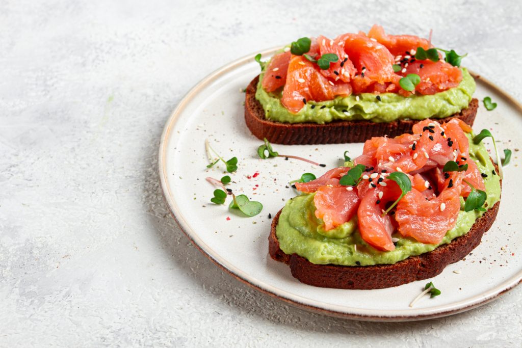Zwei Avocado-Toasts mit Räucherlachs, Sesam und Microgreens auf einem Teller.