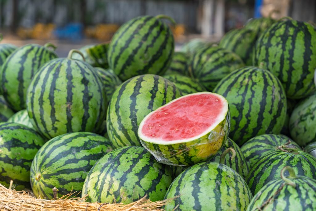 Mehrere ganze Wassermelonen mit einer aufgeschnittenen Melone, die das rote, saftige Fruchtfleisch zeigt.