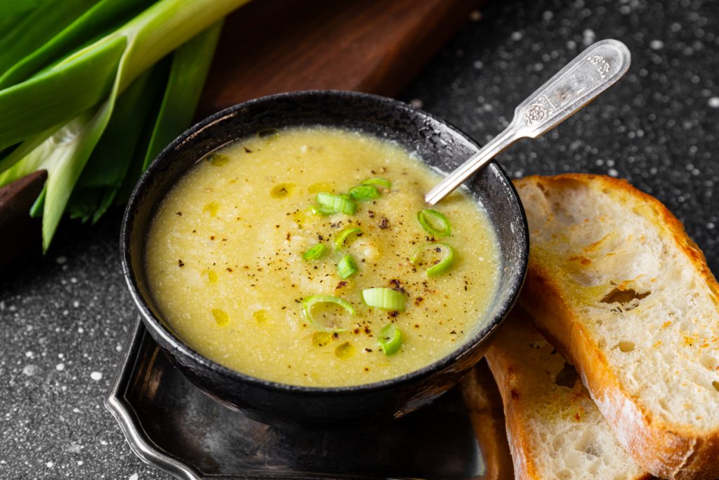 Schwarze Schale mit cremiger Lauch-Kartoffelsuppe, garniert mit Frühlingszwiebelringen, Olivenöl und schwarzem Pfeffer; daneben zwei geröstete Baguettescheiben und ein Löffel.