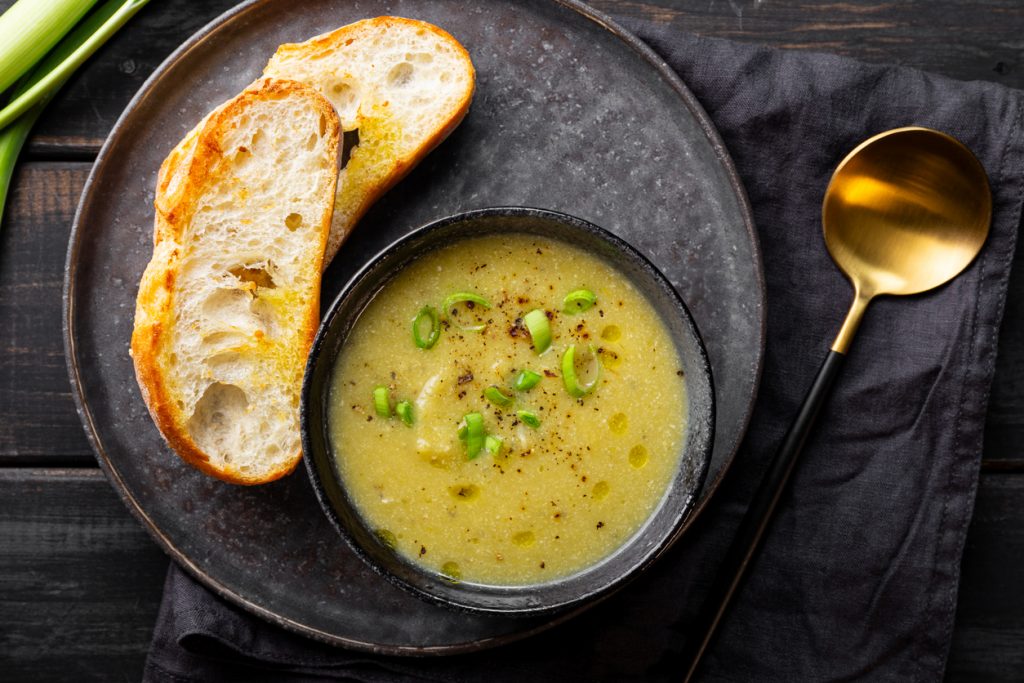 Draufsicht auf eine Schale Lauch-Kartoffelsuppe, mit Frühlingszwiebelringen, Olivenöltröpfchen und Pfeffer; daneben zwei geröstete Baguettescheiben auf dunklem Teller mit goldenem Löffel.