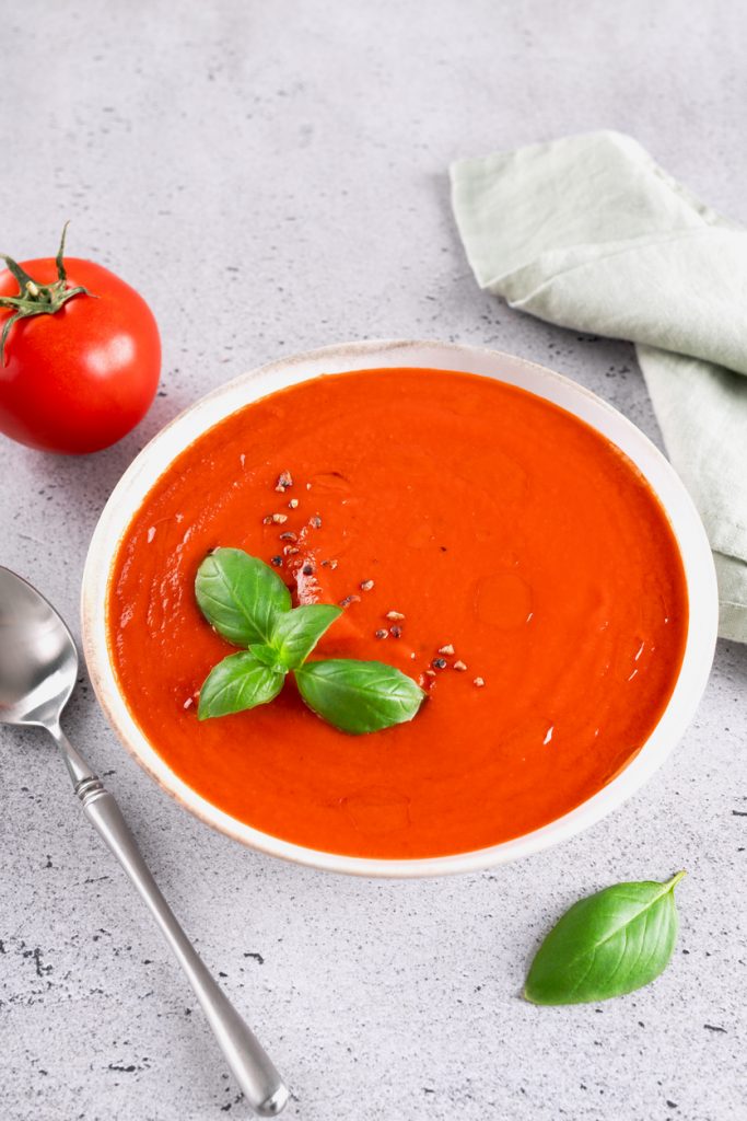 Cremige Tomatensuppe in einer weißen Schüssel, garniert mit frischem Basilikum und grobem Pfeffer, daneben eine Tomate, Basilikumblatt und ein Löffel.