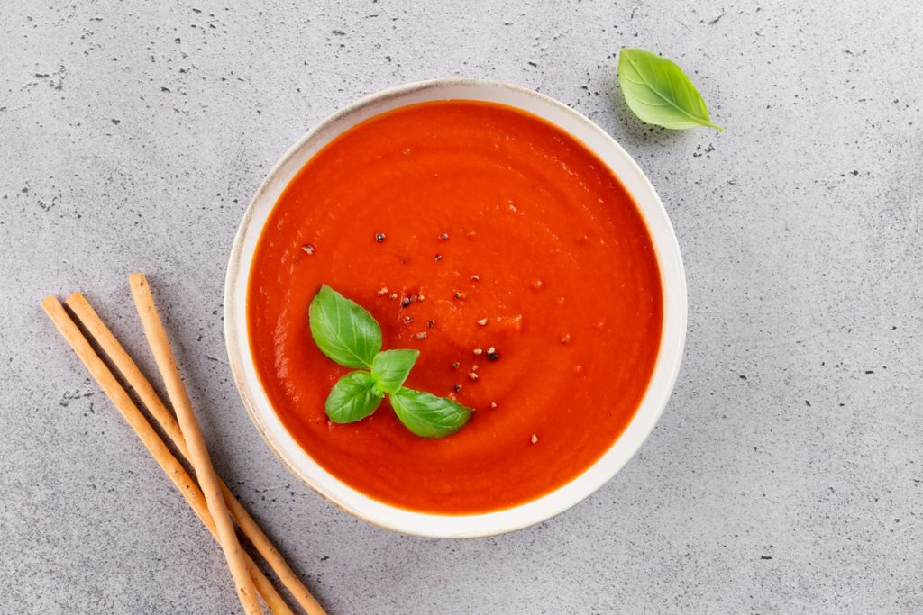 Schüssel mit cremiger Tomatensuppe, garniert mit Basilikumblättern und grobem Pfeffer, daneben Grissini-Stangen auf grauem Untergrund.