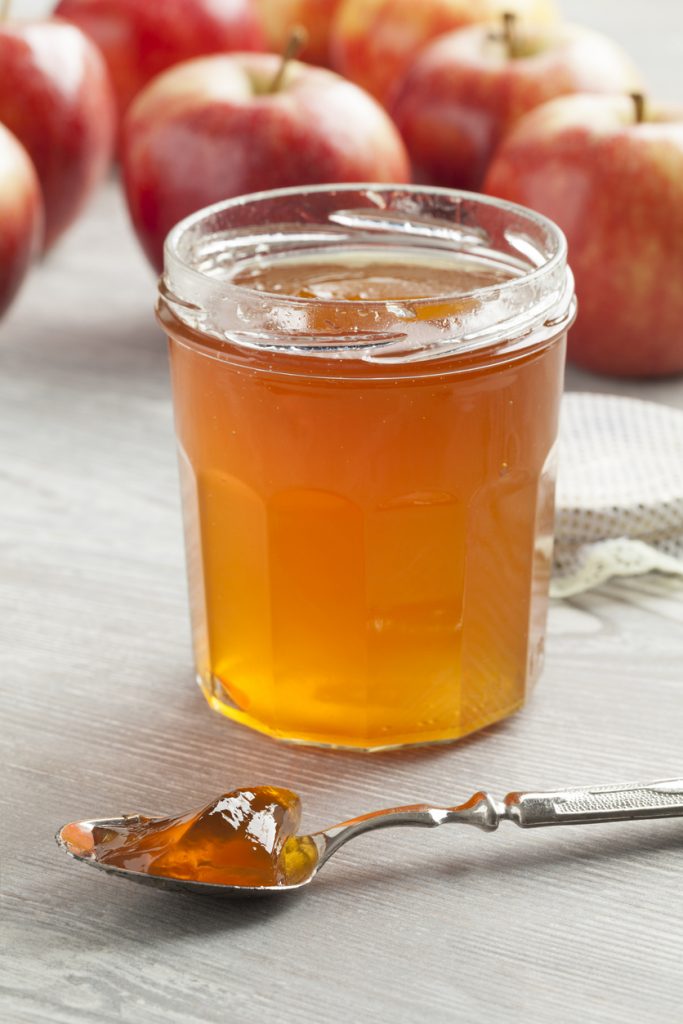 Glas mit bernsteinfarbenem Apfelgelee und Löffel, rote Äpfel im Hintergrund.