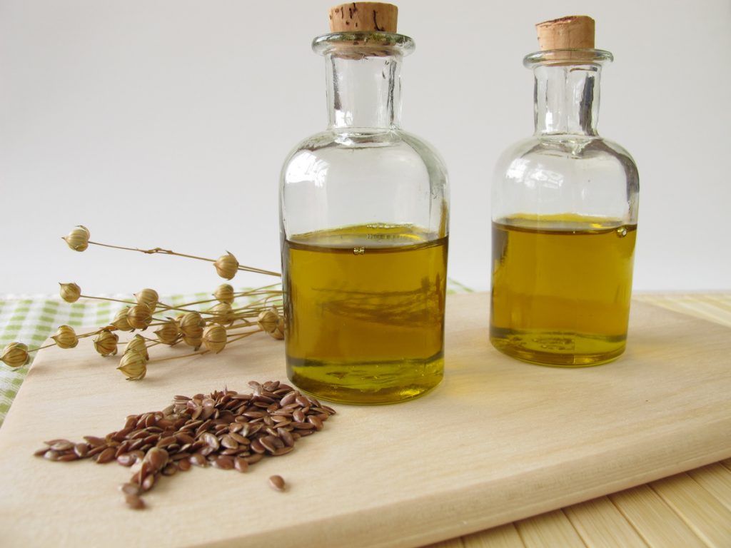 Zwei kleine Glasflaschen mit goldenem Leinöl, davor braune Leinsamen und getrocknete Flachskapseln auf einem hellen Holzbrett vor neutralem Hintergrund.