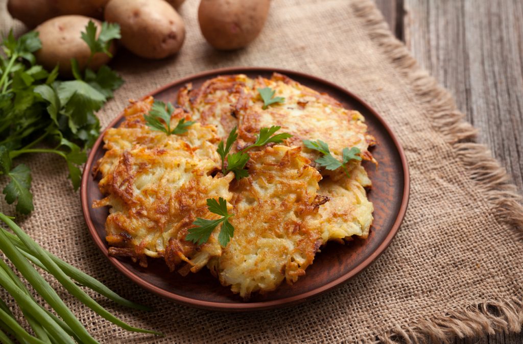 Mehrere goldbraune Kartoffelpuffer auf einem braunen Teller, mit Petersilie garniert; dahinter rohe Kartoffeln, Frühlingszwiebeln und Kräuter auf Jutesack und Holztisch.