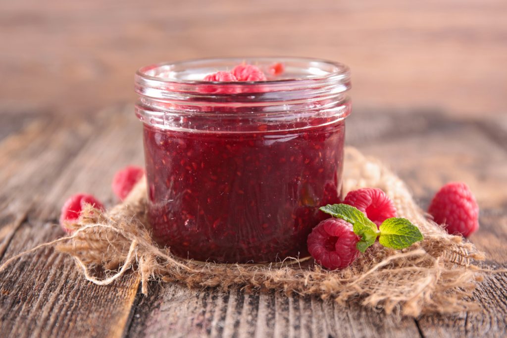 Glas Himbeermarmelade auf einem Holztisch, dekoriert mit frischen Himbeeren und Minzblättern.