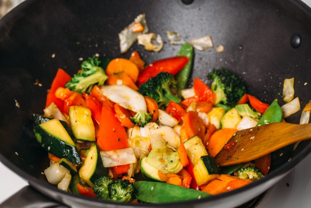 Buntes Wokgemüse mit Zucchini, Paprika, Karotten und Brokkoli in schwarzer Pfanne, Holzspatel daneben.