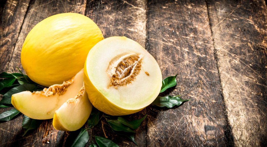 Ganze und aufgeschnittene Honigmelone mit hellem Fruchtfleisch und Kernen auf rustikalem Holztisch angerichtet.