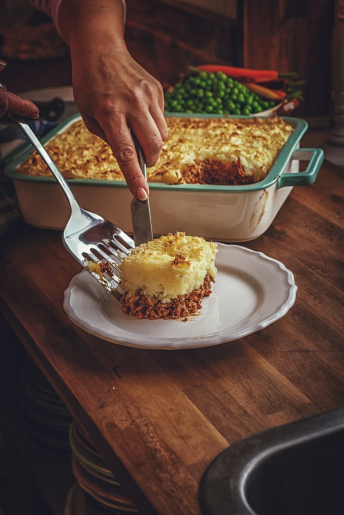Hand setzt ein Stück Shepherd’s Pie mit Kartoffelpüreehaube aus der Auflaufform auf einen Teller.
