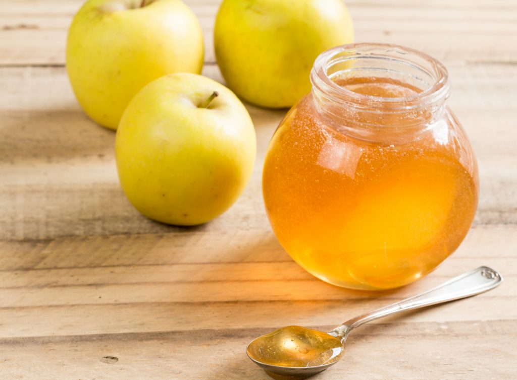 Glas mit goldgelbem Apfelgelee, gelbe Äpfel und Löffel auf Holztisch.