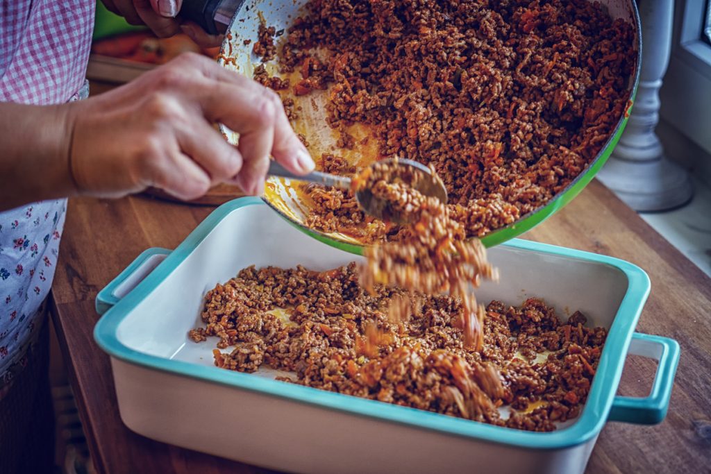 Hand füllt angebratenes Hackfleisch aus der Pfanne in eine rechteckige Auflaufform für Shepherd’s Pie.