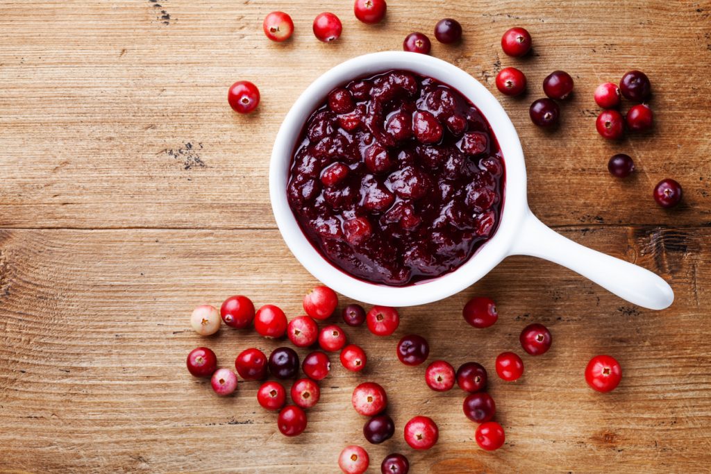 Draufsicht auf weiße Schale mit stückiger Cranberrysauce und verstreuten Cranberries auf Holztisch.