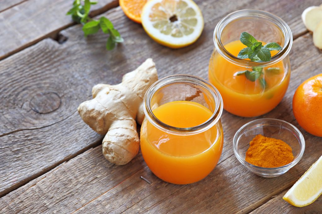 Zwei kleine Gläser mit leuchtend orangem Ingwershot, eines mit Minze garniert; daneben Ingwerwurzel, Schälchen mit Kurkumapulver, Zitronen- und Orangenscheiben auf rustikalem Holztisch.