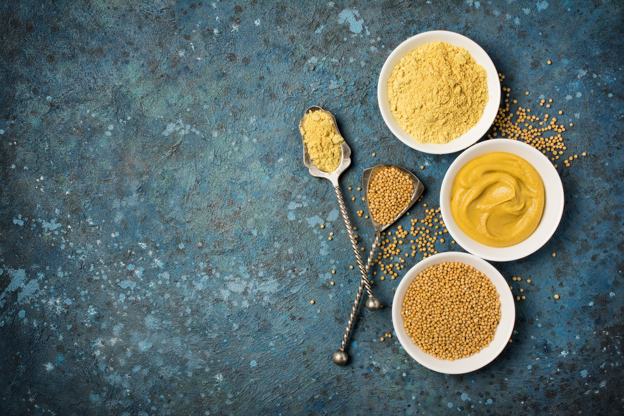 Schalen und Löffel mit Senfkörnern, Senfmehl und cremigem Senf auf blauem Untergrund.