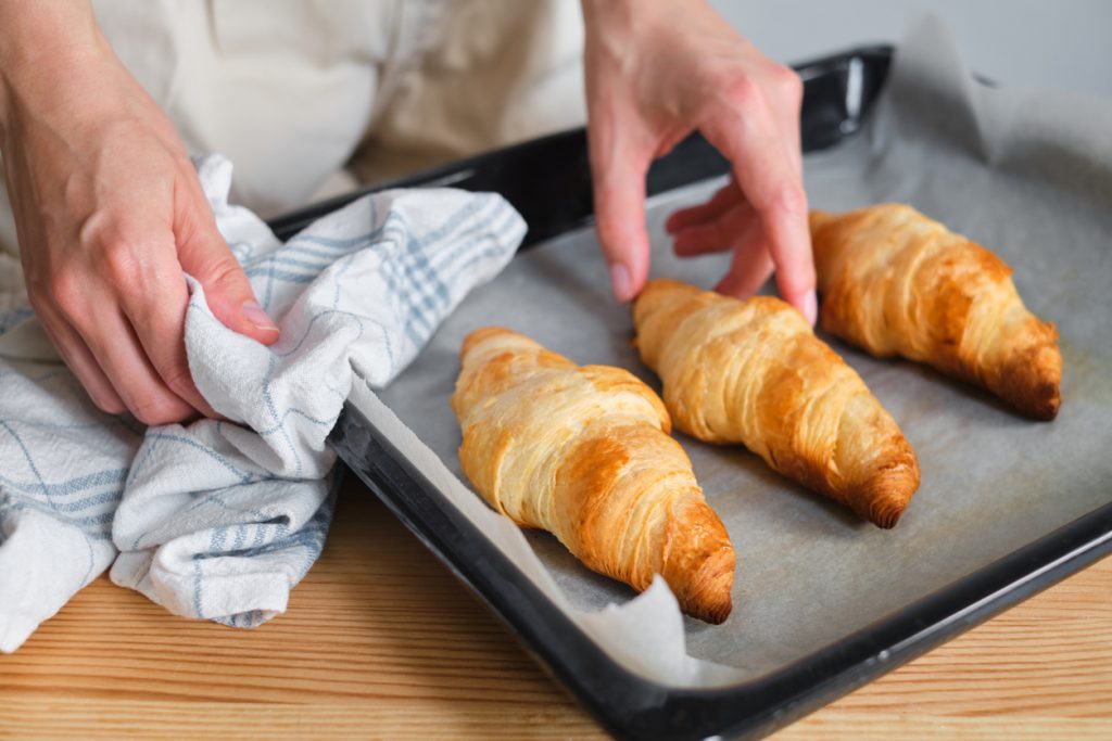 Hand richtet drei goldbraune Buttercroissants auf einem mit Backpapier belegten Blech, gehalten mit einem Küchentuch.