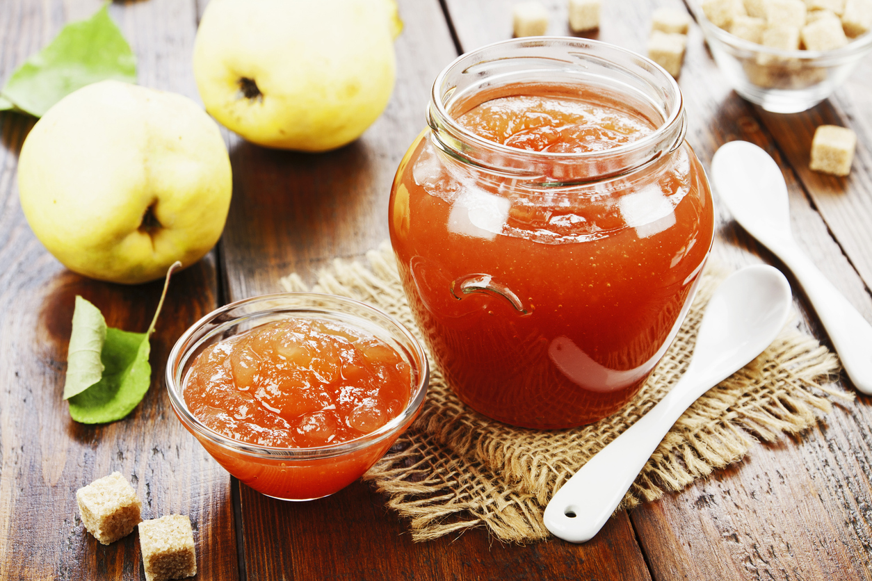 Großes Glas und kleine Schale mit orangefarbener Quittenkonfitüre auf Holztisch, umgeben von Quitten, Zuckerwürfeln und zwei weißen Löffeln.