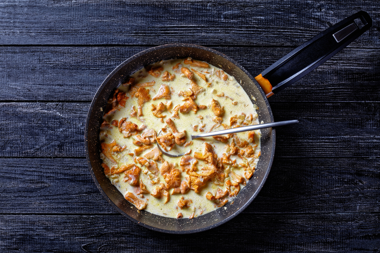 Draufsicht auf eine Pfanne mit heller Pfifferlingssauce, orange Pilzstücken und einem Löffel auf dunklem Holztisch.