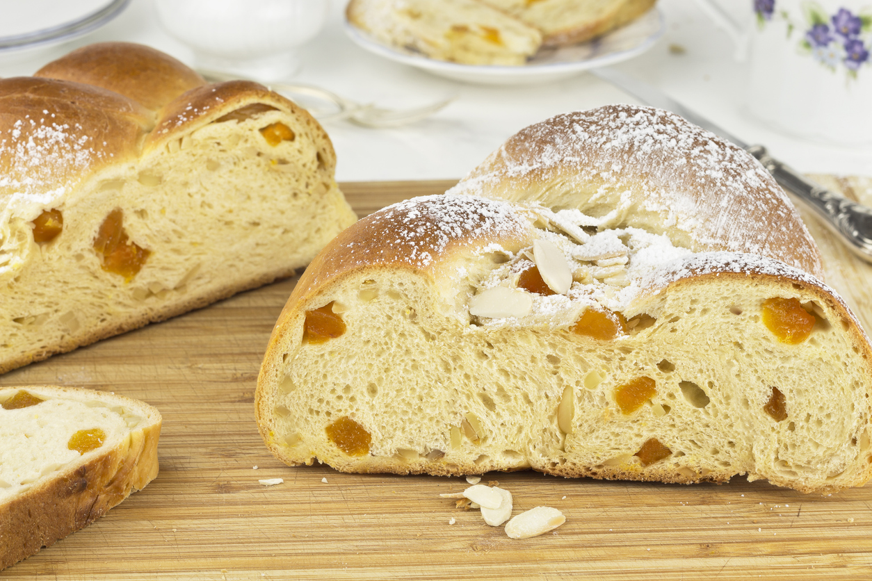 Ein goldbrauner Hefezopf mit Aprikosenstücken und Mandelblättchen liegt auf einem Holzbrett und ist angeschnitten.