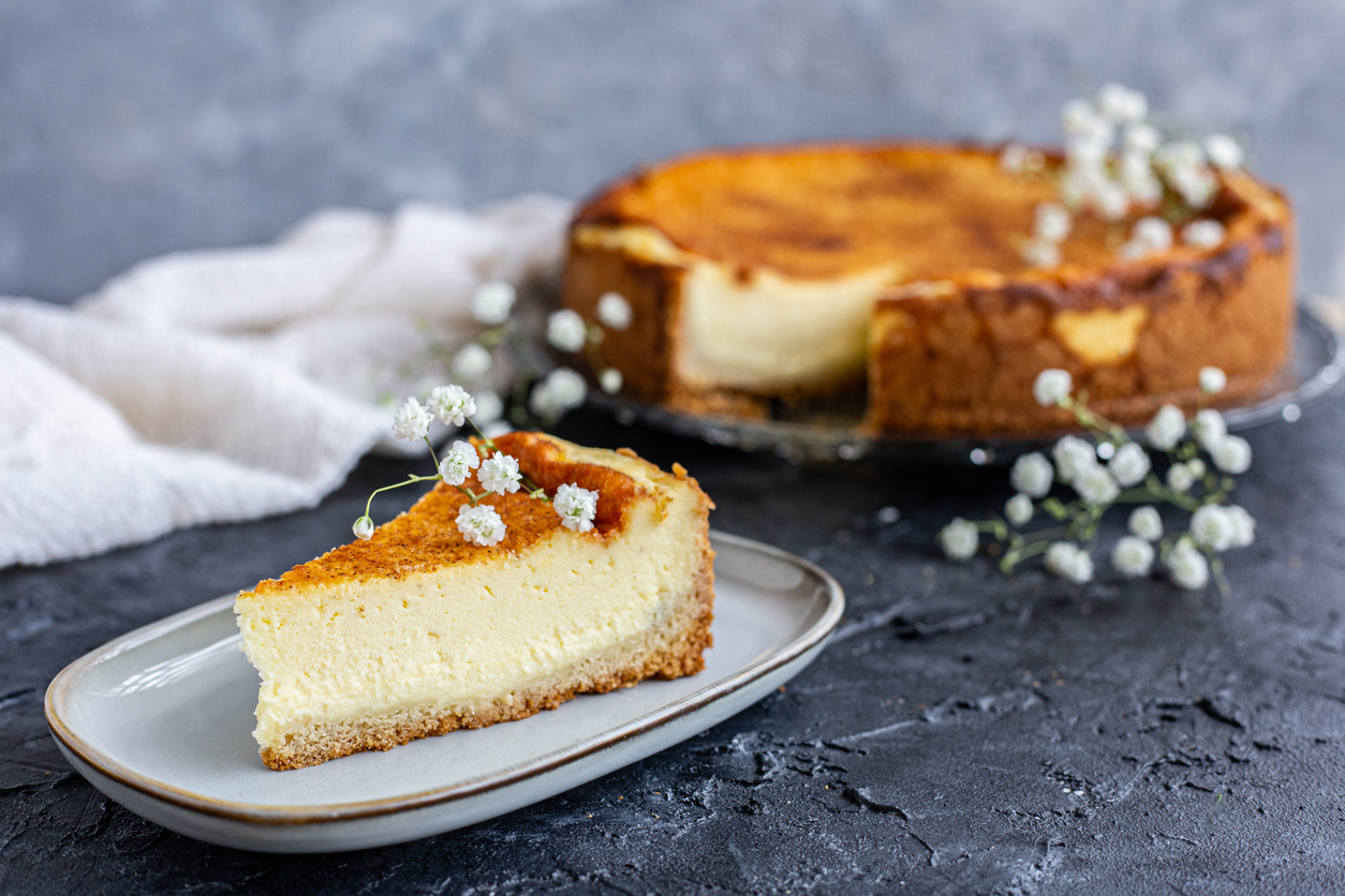 Ein Stück cremiger Käsekuchen auf einem Teller, dahinter der angeschnittene Kuchen auf einer Platte mit kleinen weißen Blüten.
