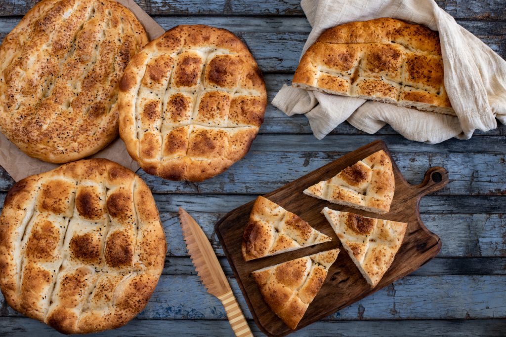 Goldbraune Pitabrote mit Rautenmustern, teils angeschnitten auf einem Holzbrett neben Brotmesser.